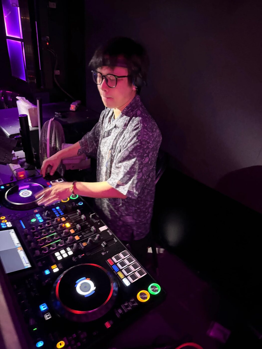 DJ 在活動現場的控制器前混歌，雙手放在播放轉盤與混音推桿上，旁邊是自帶的器材與電源線