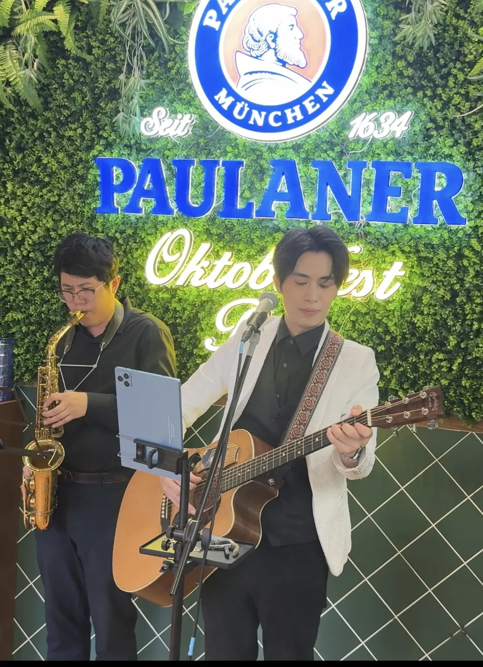 品牌活動實景:綠色植物牆前,一位樂手吹奏 Saxophone,另一位穿白色外套的樂手彈木結他並唱歌,面前是一支咪高峰與夾著平板電腦的譜架